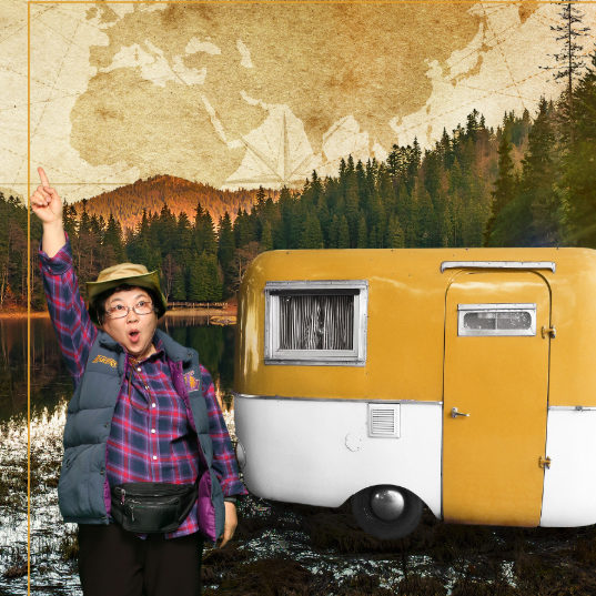 Jeongsun standing next to a vintage caravan, pointing to the sky, with ranges of pine trees in the distance
