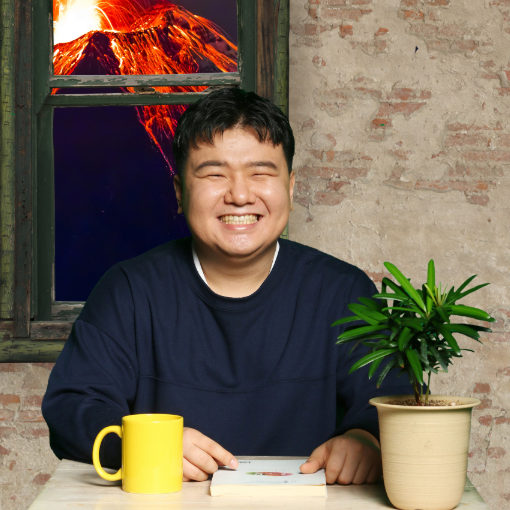 Yongin sitting and smiling at a table. Through the window behind him, a volcano is erupting outside.