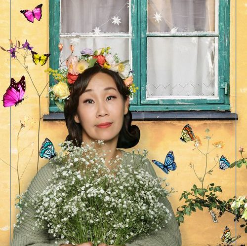 Sujin wears a floral wreath on her head and holds a bunch of white flowers surrounded by butterflies. 